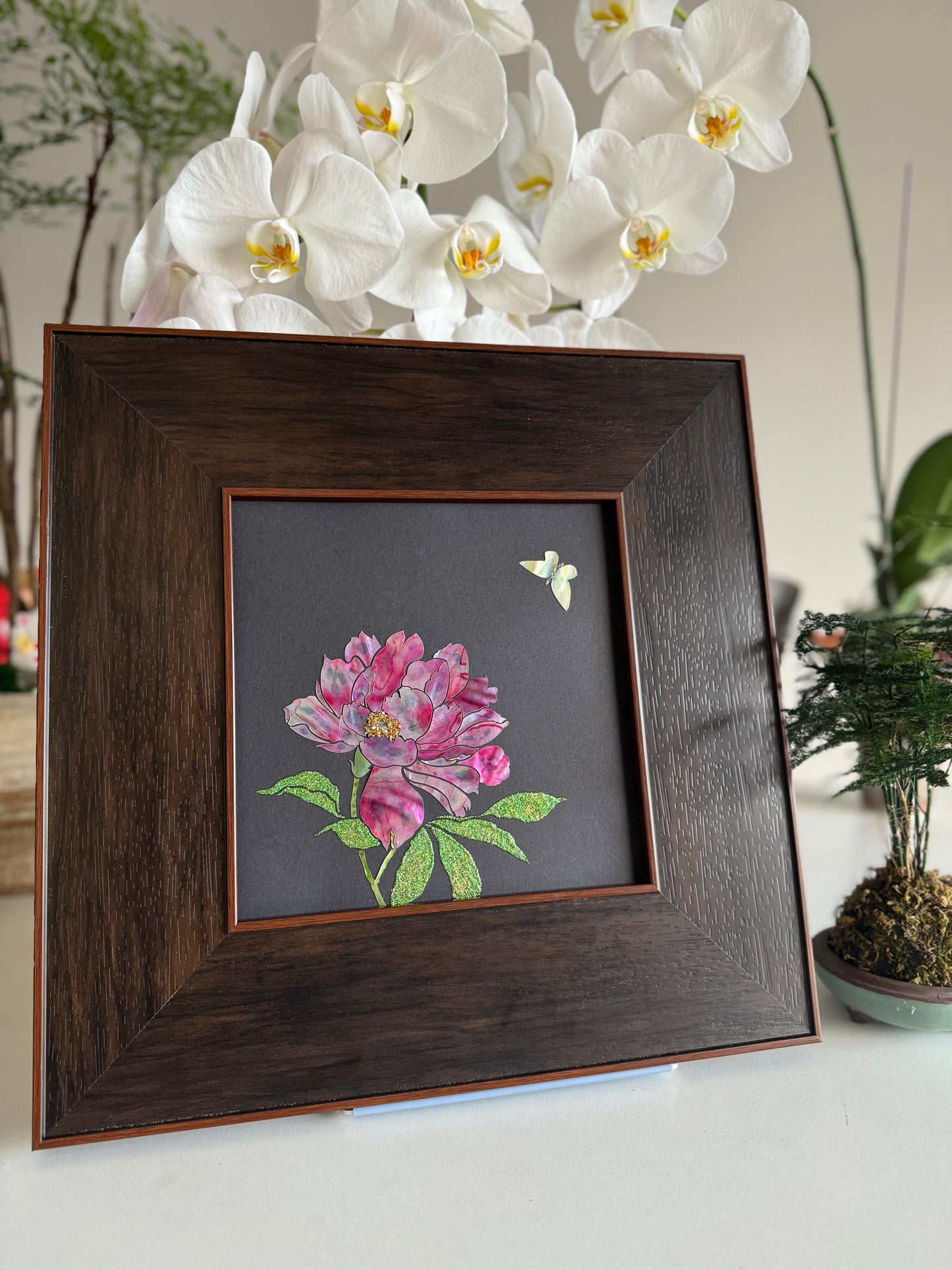 Premium boutique botanical wall decor displaying the handcrafted "Blossom of Radiance" shell inlay artwork on a desktop near white orchids and a miniature green plant.