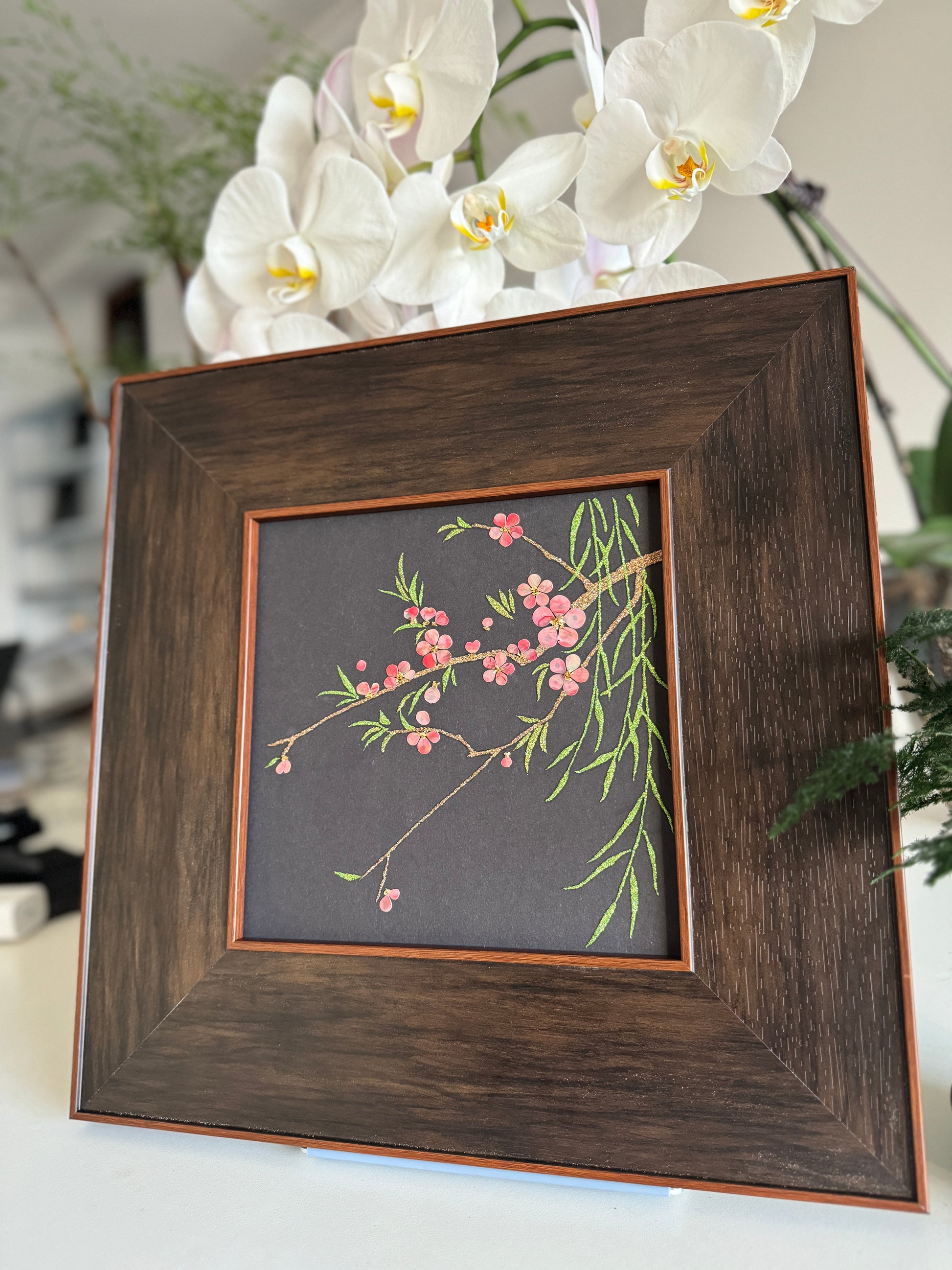 Premium boutique botanical home decor presentation featuring the handcrafted "Smile of Spring" peach blossom shell mosaic painting displayed against a backdrop of elegant white orchids.