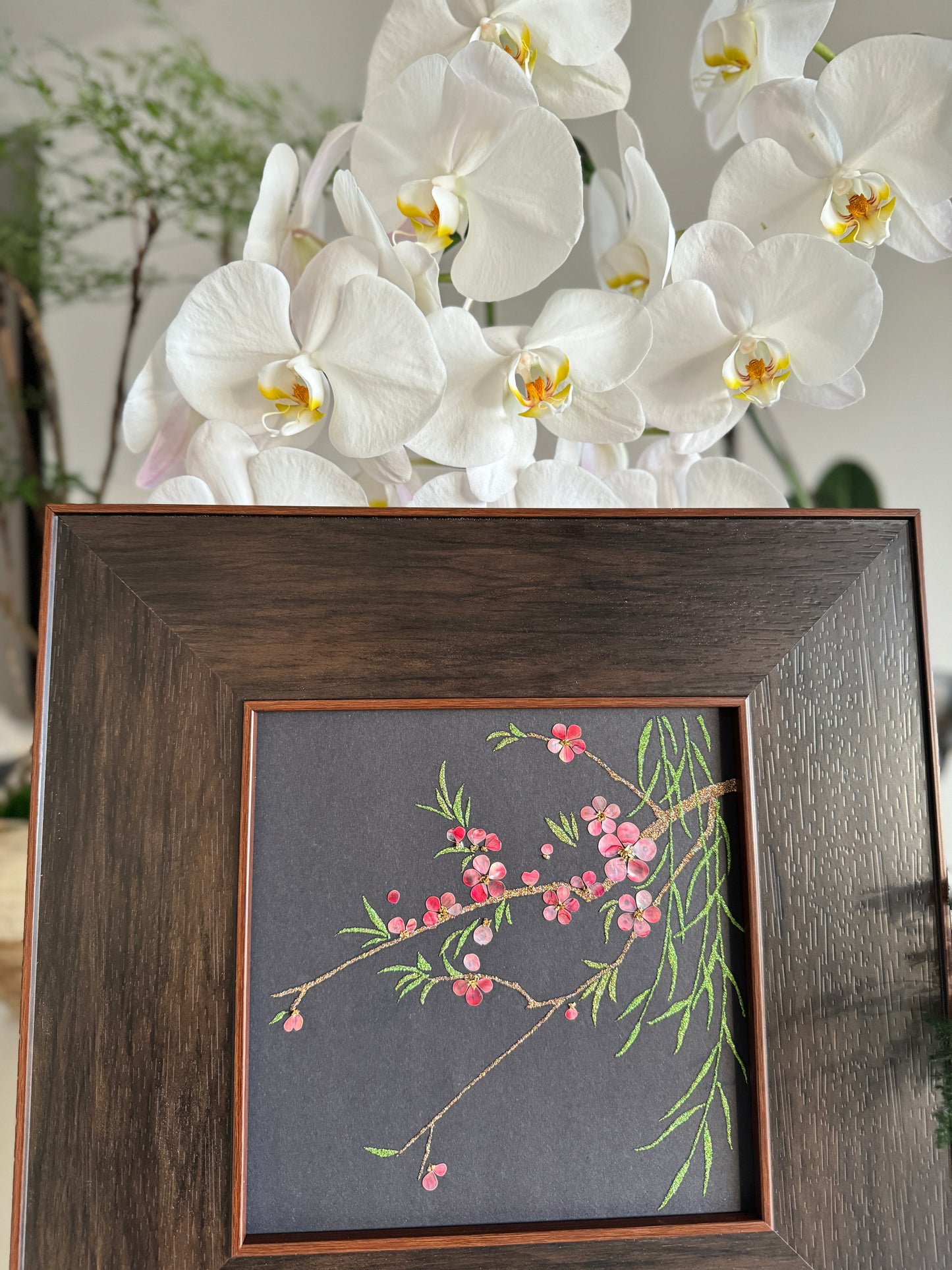 Premium boutique botanical home decor presentation featuring the handcrafted "Smile of Spring" peach blossom shell mosaic painting displayed against a backdrop of elegant white orchids.