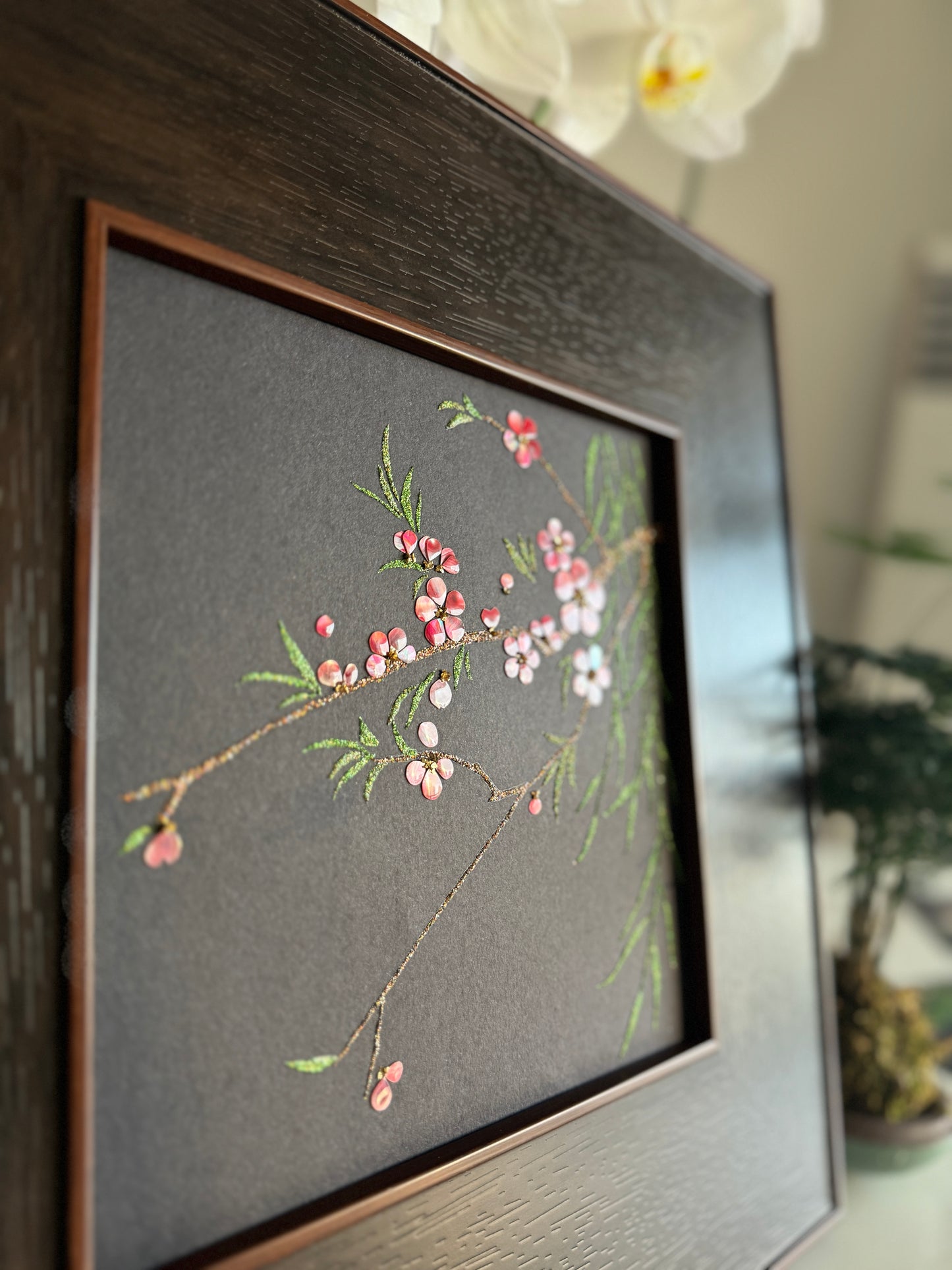 Premium boutique botanical home decor presentation featuring the handcrafted "Smile of Spring" peach blossom shell mosaic painting displayed against a backdrop of elegant white orchids.