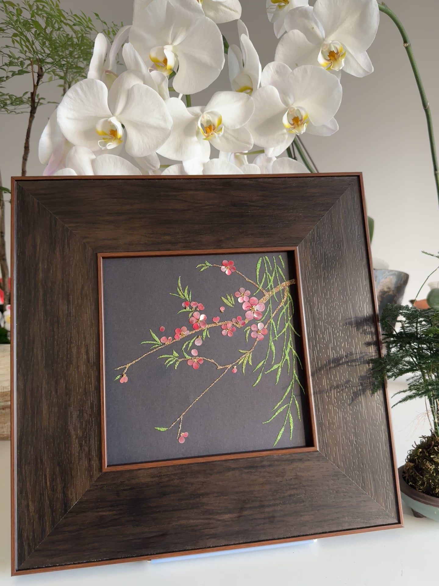 Premium boutique botanical home decor presentation featuring the handcrafted "Smile of Spring" peach blossom shell mosaic painting displayed against a backdrop of elegant white orchids.